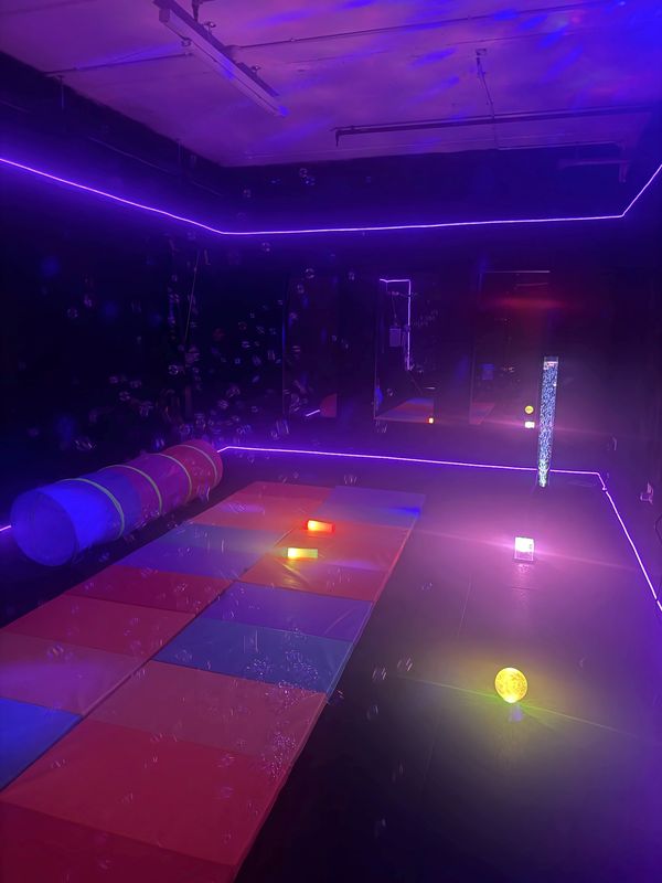 Sensory room bathed in purple light, with colourful soft mats, a play tunnel, a bubble tube and floating bubbles