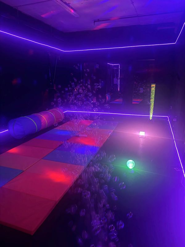 Bubbles drifting over rainbow soft mats in the purple-lit sensory room, with a light-up ball glowing on the floor